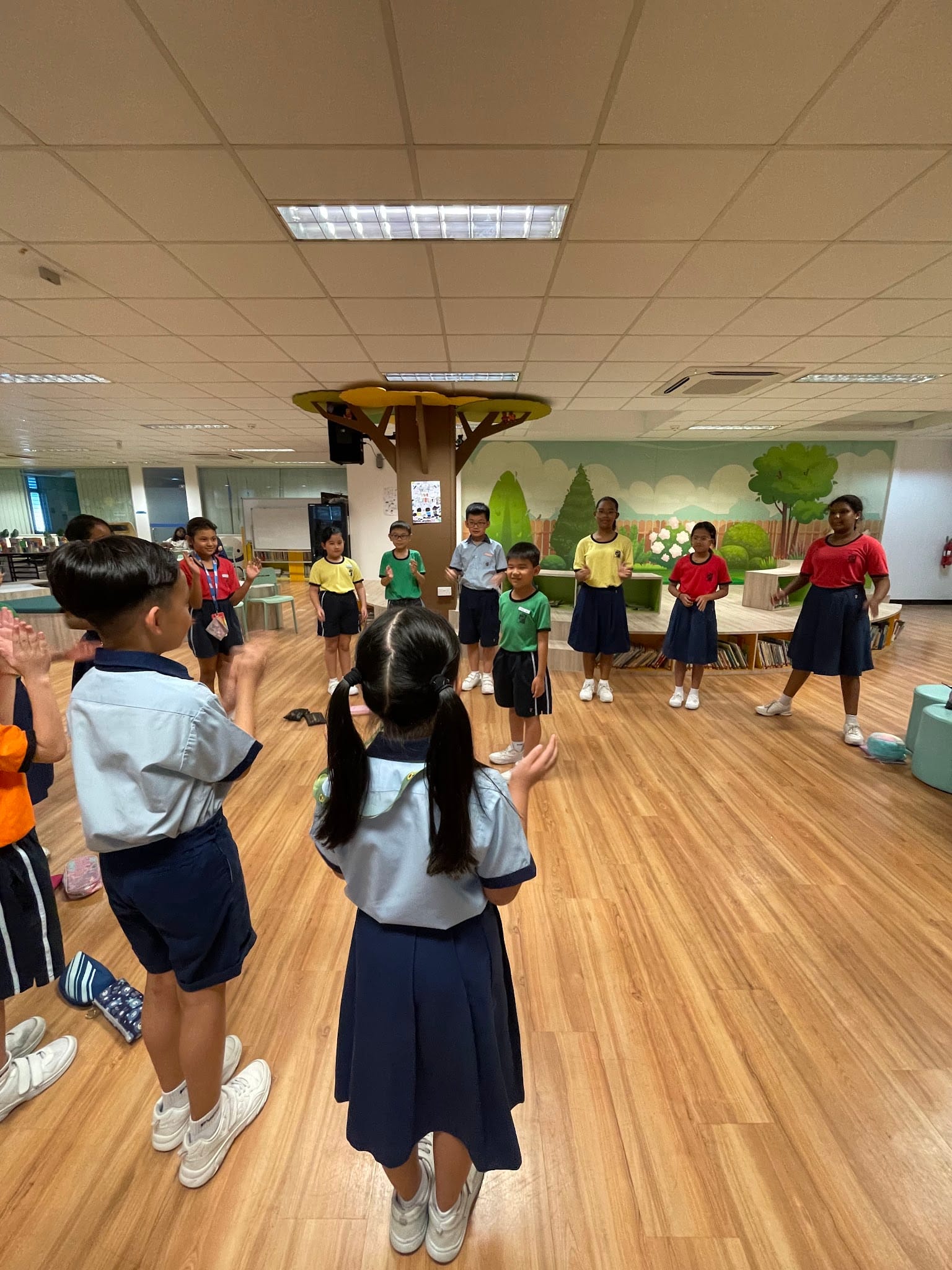 A group of primary school children in colorful uniforms standing in a circle and clapping during an activity in a wooden-floored room.